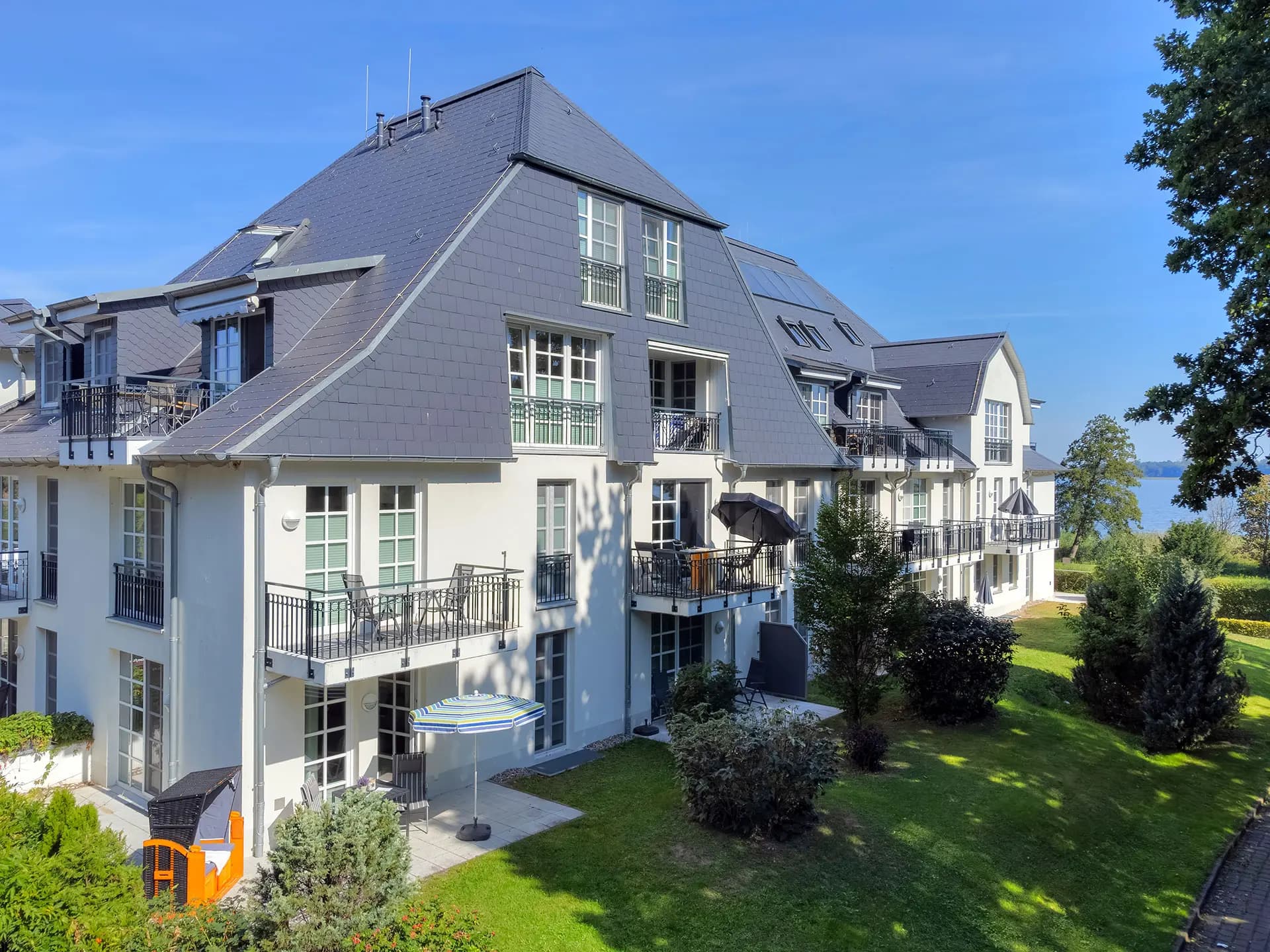 Biały budynek apartamentowy z balkonami i łupkowym dachem w otoczeniu zieleni